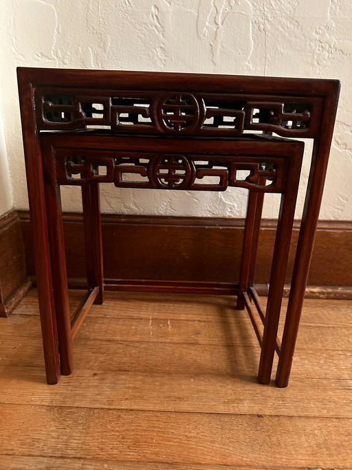 A set of two intricately carved wooden nesting tables features a rich mahogany finish and decorative openwork designs, resting on a wooden floor.