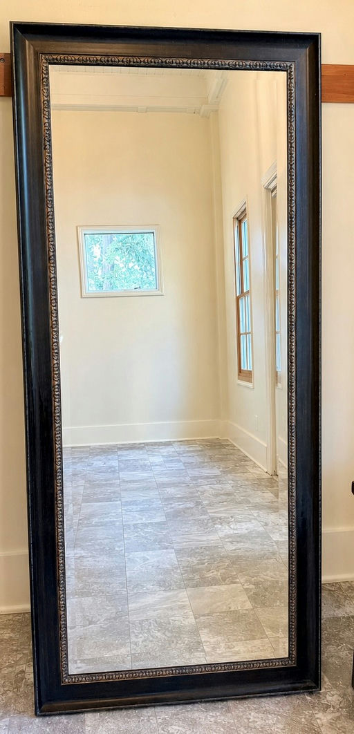 A tall, rectangular mirror with a decorative black frame reflects a bright, empty room featuring a gray tiled floor and white walls.