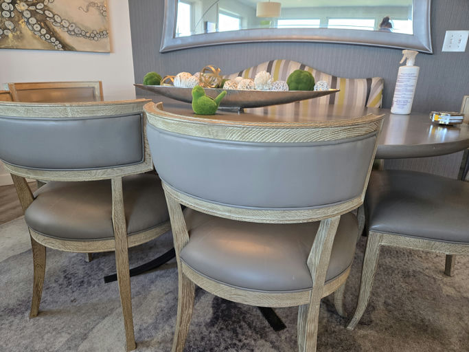 A dining area features elegant wooden chairs with gray upholstered seats, a round table, and decorative elements on the tabletop. Natural light filters in.