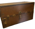 A vintage brown trunk with metal latches sits on an ornate, colorful rug. The wall behind is a plain light color, creating a simple backdrop.