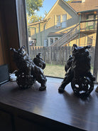 Two decorative lion sculptures sit on a windowsill, framed by sunlight, overlooking a yard and a house with a staircase in the background.