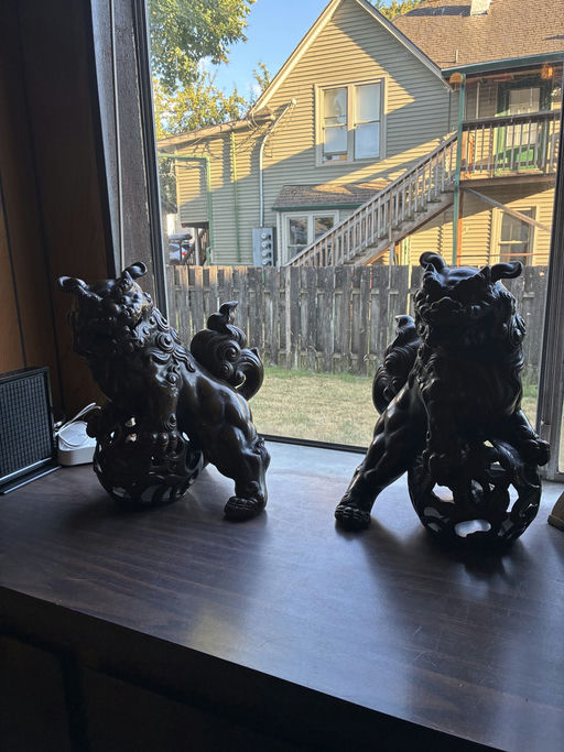 Two decorative lion sculptures sit on a windowsill, framed by sunlight, overlooking a yard and a house with a staircase in the background.