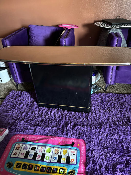 A black table with a shiny top sits in a cozy room, flanked by purple chairs and a pink play mat with colorful designs on a plush purple rug.