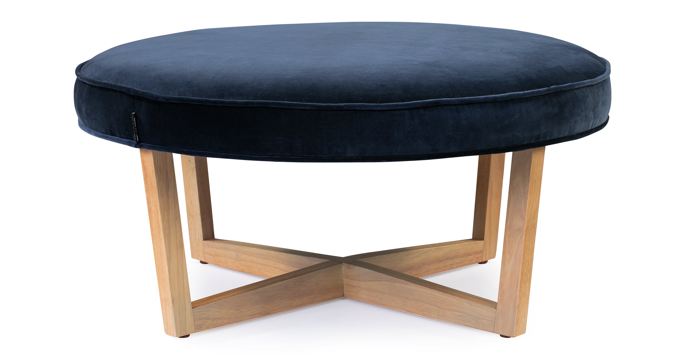 Compass ottoman has an upholstered cushioned top that is in the shape of a circle. The cushioned top has piping along the edges. There are 4 natural wood legs that angle in slightly from the top of the ottoman touch the floor. The legs connect underneath the ottoman making a shape of an X.