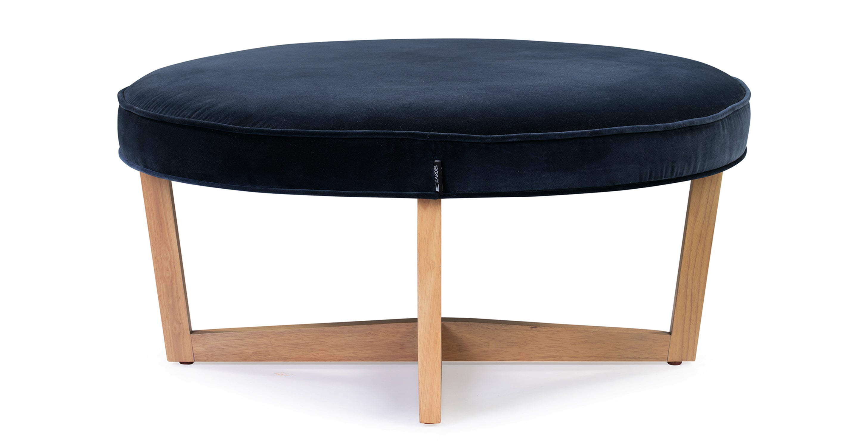 Compass ottoman has an upholstered cushioned top that is in the shape of a circle. The cushioned top has piping along the edges. There are 4 natural wood legs that angle in slightly from the top of the ottoman touch the floor. The legs connect underneath the ottoman making a shape of an X.