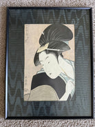 A framed ukiyo-e print features a woman with elaborate black hair and traditional clothing, holding a brush and gazing thoughtfully.