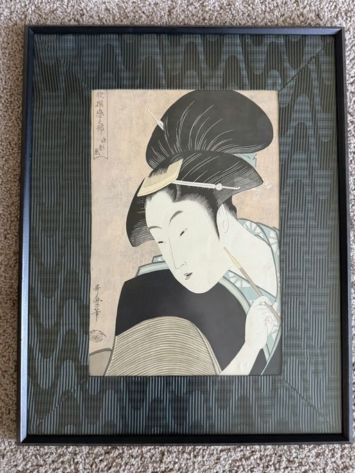 A framed ukiyo-e print features a woman with elaborate black hair and traditional clothing, holding a brush and gazing thoughtfully.