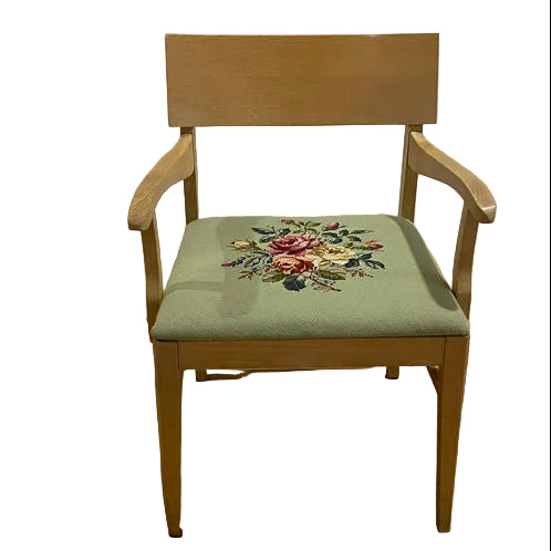 A wooden chair with armrests features a green upholstered seat adorned with floral embroidery, placed on a brown mat over hardwood flooring.