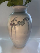 A decorative vase with a soft, speckled finish featuring elegant botanical drawings in muted colors, accented by green leaves peeking out.