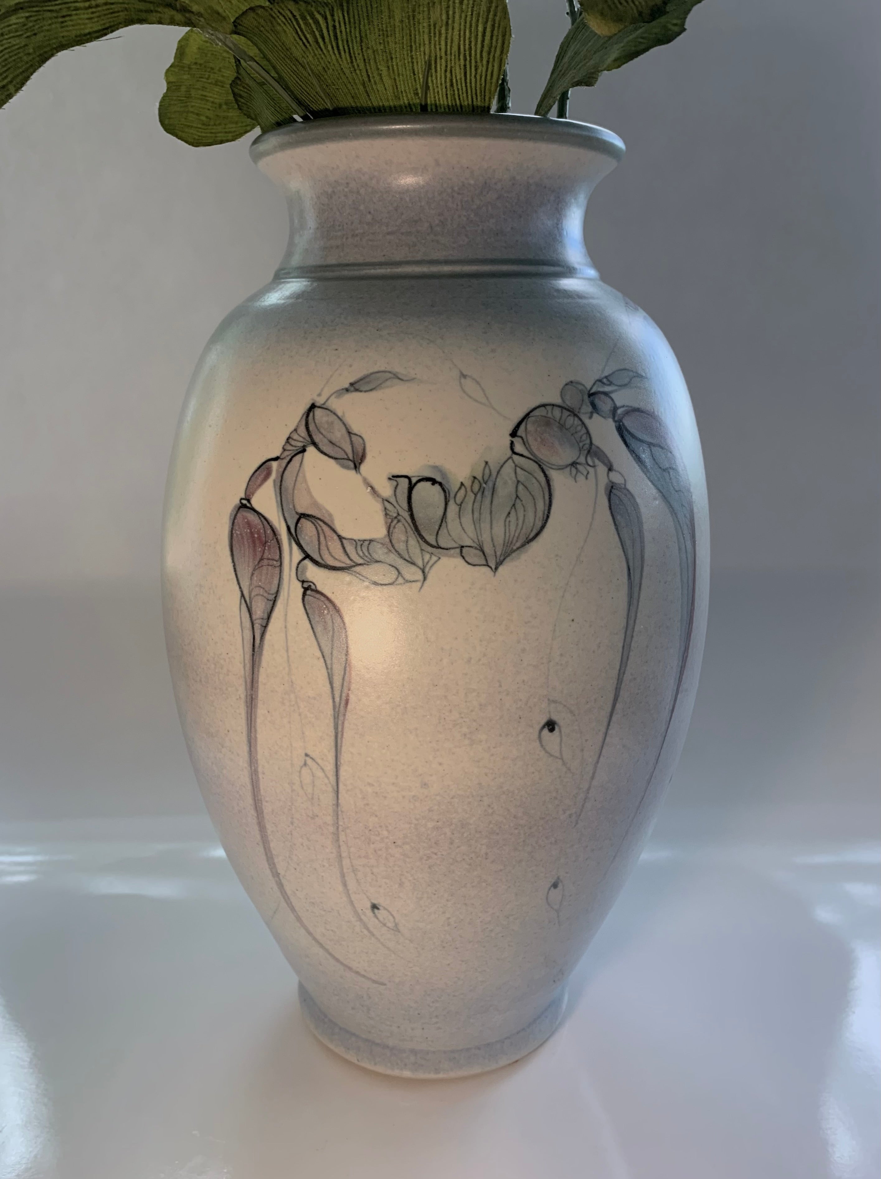 A decorative vase with a soft, speckled finish featuring elegant botanical drawings in muted colors, accented by green leaves peeking out.