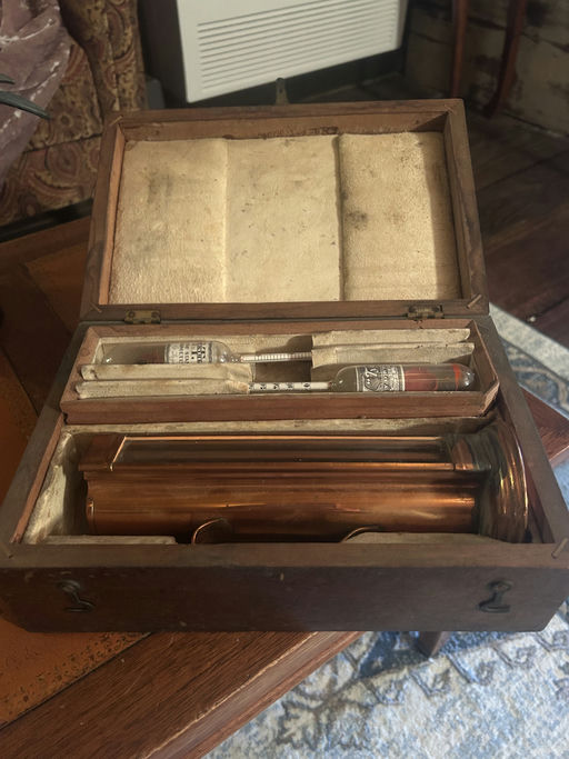 A vintage wooden box is open, displaying a copper instrument, glass vials, and other components, all resting on a felt-lined interior.