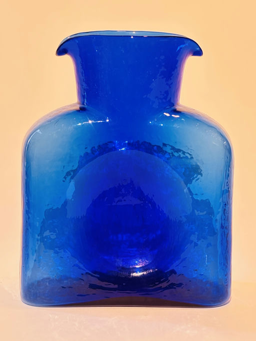 A vibrant blue glass vase with a unique shape, featuring a wide body and a narrow spout, placed against a soft, neutral background.