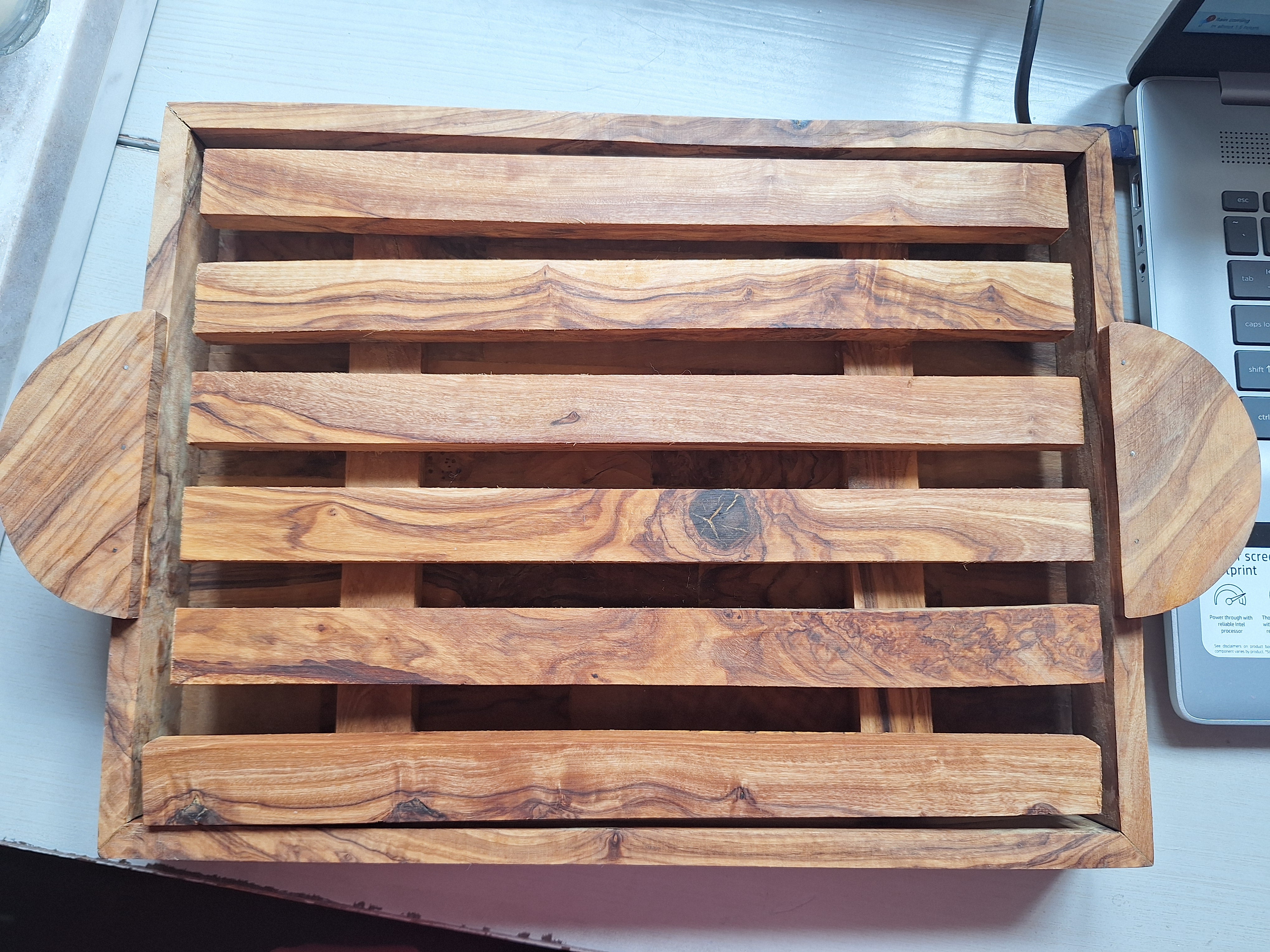 A wooden serving tray with slatted design and rounded handles, resting on a flat surface next to a laptop. Natural grain patterns visible.
