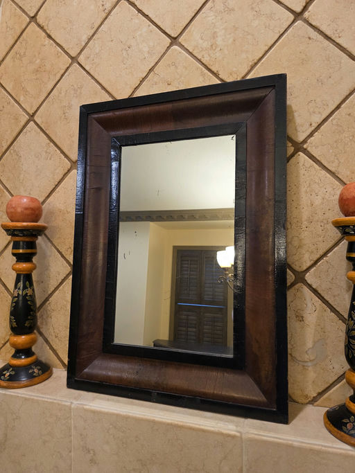 A rectangular mirror bordered with dark wood is mounted on a tiled wall. Two decorative candlesticks are positioned on either side of the mirror.