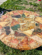 A close-up of a colorful mosaic table, featuring various polished stones in hues of orange, red, green, and tan, set against green grass.