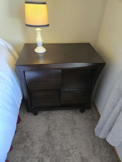 A dark wooden nightstand with two square compartments sits beside a bed. On top, there's a white lamp with a yellow shade, illuminating the space.
