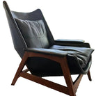A sleek black leather chair with wooden legs sits beside a window, casting soft light across a striped rug and illuminating the cozy interior.