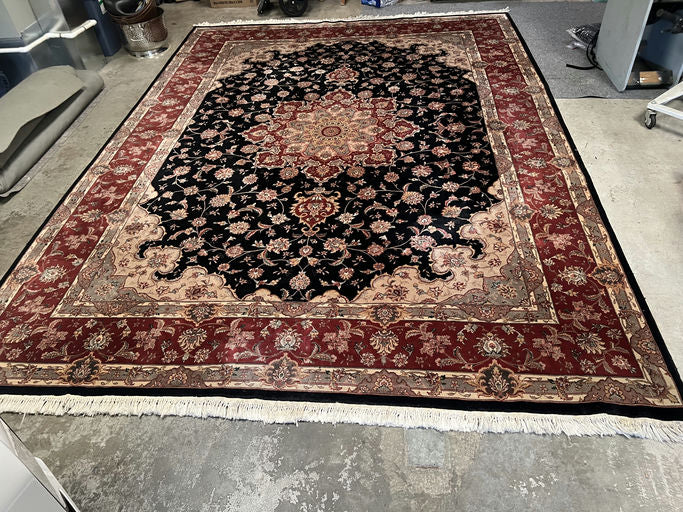 A large, intricate Persian rug featuring a floral pattern in rich black, red, and gold tones, laid on a concrete floor.
