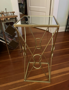 A gold metal side table with a glass top. It features an intricate design with curved elements and stands on a wooden floor, against a light wall.