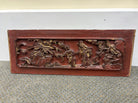 A decorative wooden panel featuring intricate relief carvings of flowers and figures, painted in red and gold, resting on a carpeted floor.