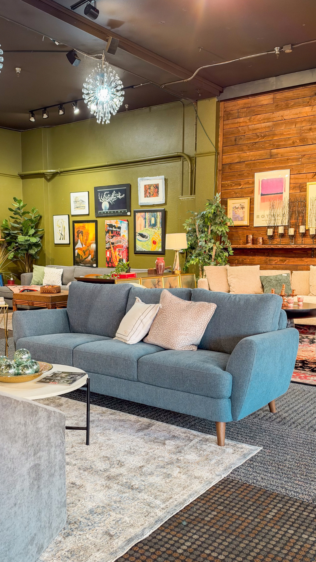 A blue upholstered sofa with light beige and white throw pillows, positioned in a styled living room showroom with a wooden wall, framed art, and other furniture in the background.