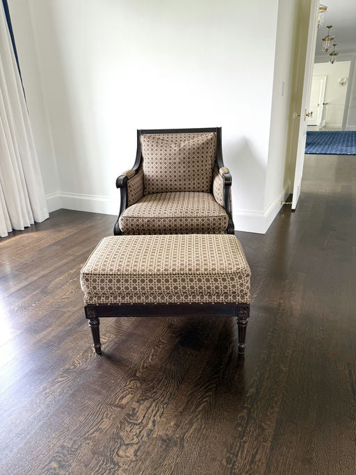 A vintage upholstered chair with an ottoman sits on dark hardwood flooring in a well-lit room with white walls and neutral decor.