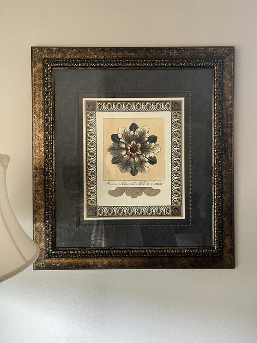 A framed botanical print featuring a floral design, accented with ornate borders and a decorative background, hung on a wall near a lamp.