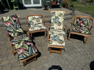 Four wooden patio chairs with floral and palm-patterned cushions sit on a stone patio, with greenery and furniture in the background.