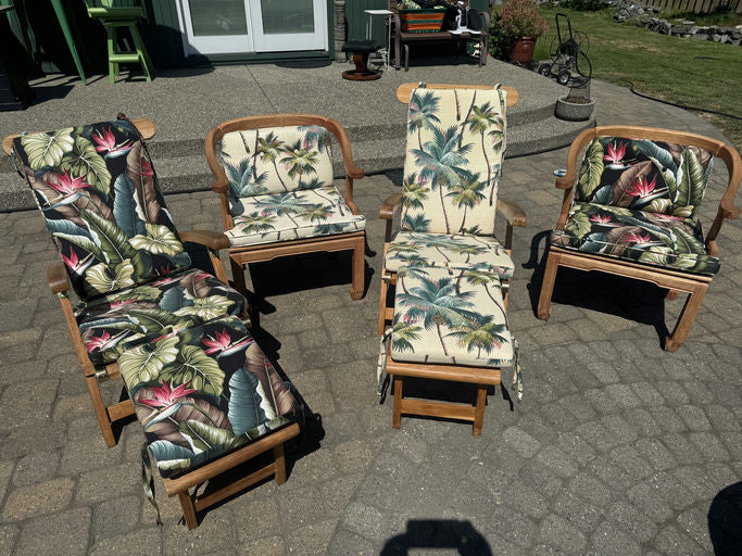 Four wooden patio chairs with floral and palm-patterned cushions sit on a stone patio, with greenery and furniture in the background.
