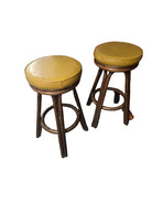 Two vintage wooden bar stools with yellow padded seats, positioned on a concrete floor. A background includes wooden furniture and a storage area.