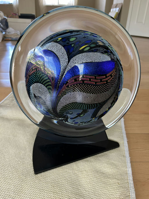 A colorful glass orb featuring intricate patterns of blue, green, and metallic accents, displayed on a sleek black stand.