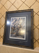 A framed artwork depicting a lighthouse, pelicans, and other birds amidst a dramatic sky, hanging on a textured tiled wall.