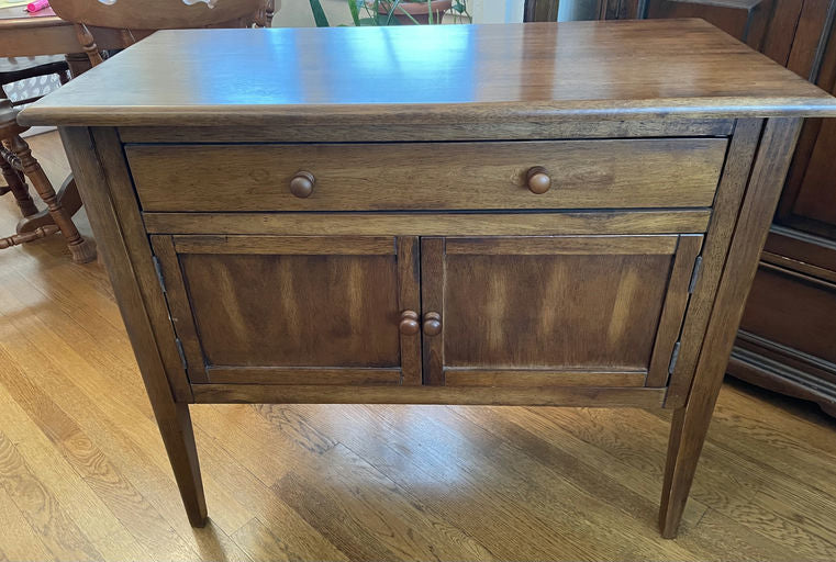 A wooden side table with a smooth surface, one drawer, and two cabinet doors, resting on four tapered legs against a hardwood floor backdrop.