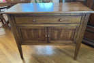 A wooden side table with a smooth surface, one drawer, and two cabinet doors, resting on four tapered legs against a hardwood floor backdrop.