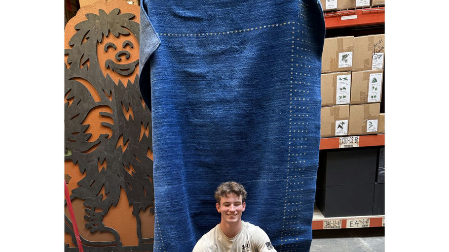 A smiling young man sits in front of a large blue fabric, next to a playful wooden figure resembling a furry creature, with shelves of boxes behind.