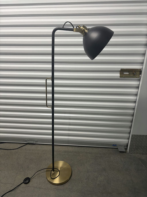 A modern floor lamp featuring a black shade and a brass accent, set against a backdrop of corrugated metal storage unit doors.