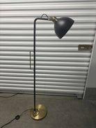 A modern floor lamp featuring a black shade and a brass accent, set against a backdrop of corrugated metal storage unit doors.