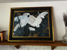 A colorful painting of two white cockatoos perched on branches, surrounded by dark green leaves, framed in an ornate black and gold frame.