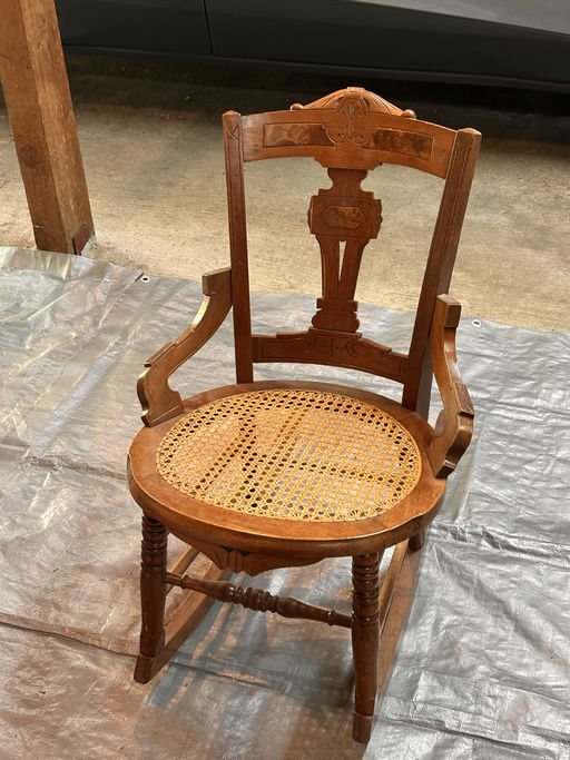 A vintage wooden chair with a woven cane seat, featuring ornate carvings on the backrest. It's placed on a tarp in a garage setting.