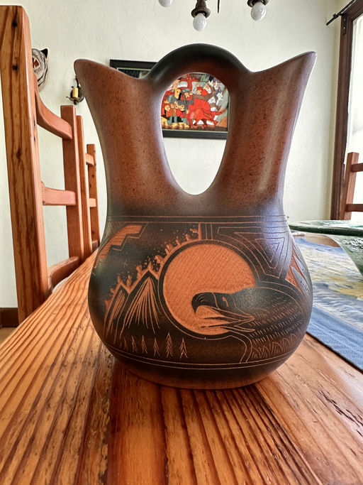 A handcrafted pottery vase with a distinctive handle, featuring intricate designs of mountains and an eagle against a reddish-brown background.