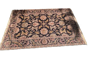 A decorative rug with intricate floral patterns in shades of black, beige, and pink placed outdoors on a textured gray surface. Shadow play is visible.