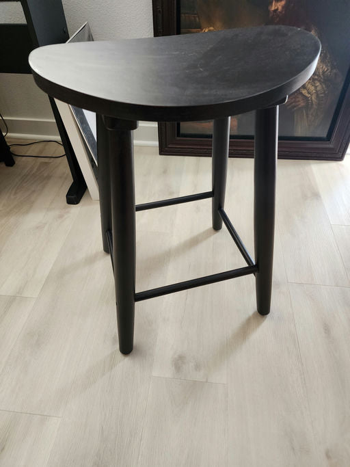 A black, half-moon shaped stool with four legs and a cross brace, positioned on light-colored flooring near framed artwork and furniture.