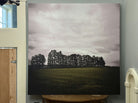 A large artwork featuring dark trees silhouetted against a cloudy sky, set above a green field. The piece is displayed on a wall near wood accents.
