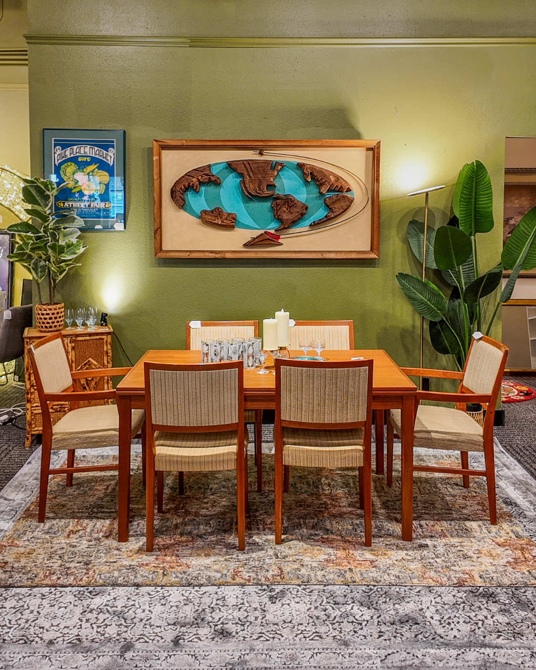A wooden dining table with six woven-back chairs is arranged on a patterned area rug in a styled room with olive green walls, wall art, plants, and warm ambient lighting.