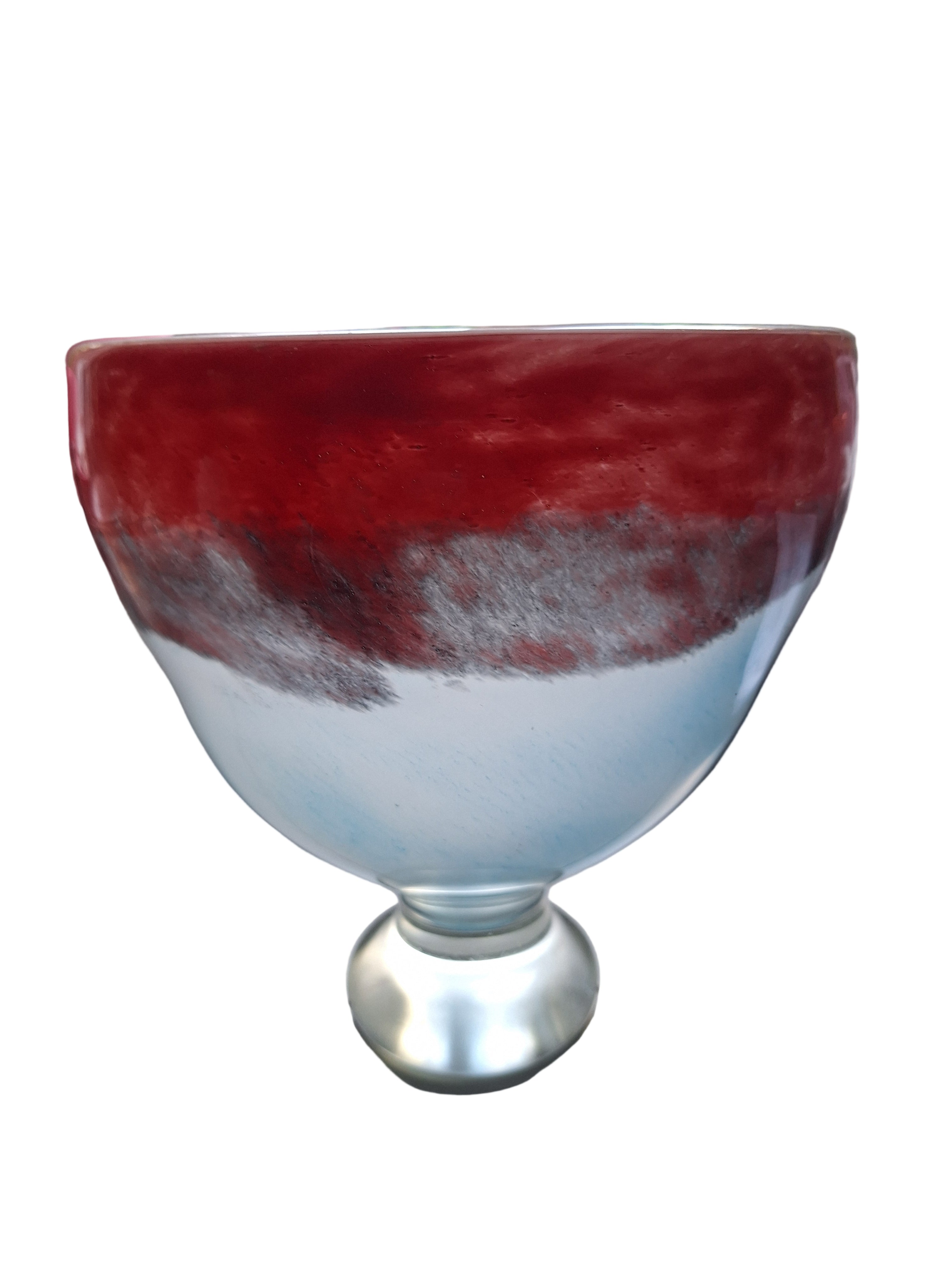 A round, decorative glass bowl featuring a red and blue gradient design, resting on a light-colored surface near a window with green foliage visible.