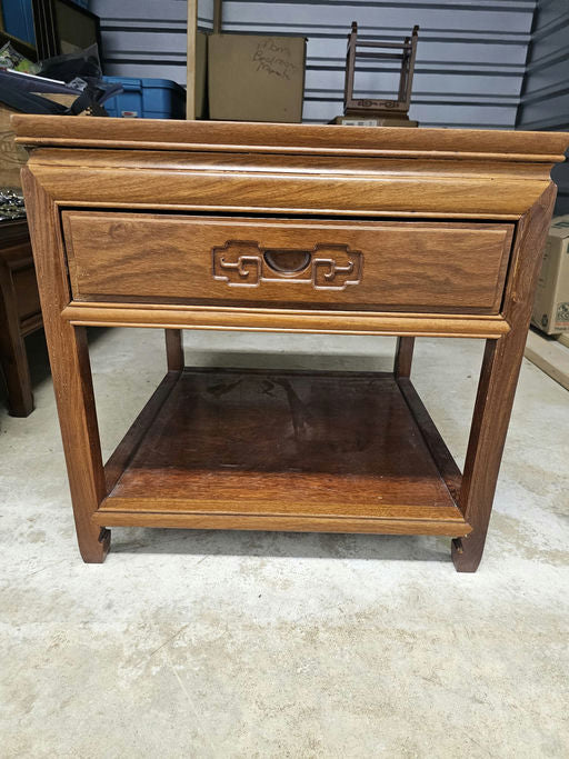 A wooden nightstand with a single drawer featuring decorative handles and an open shelf below, set against a storage unit background.