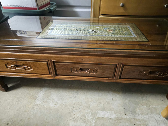 A wooden coffee table with a glass top and decorative inlay design. It features two drawers with unique handles, placed on a concrete floor.