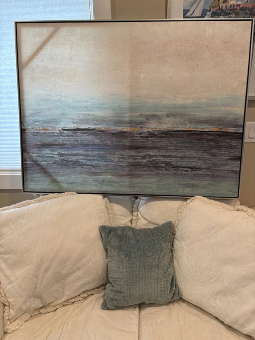 Abstract landscape painting with soft blues and browns displayed on a couch with textured white and teal pillows. Natural light from a nearby window.