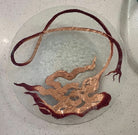 A circular glass table featuring intricate copper artwork resembling flowing waves and abstract shapes, set against a light speckled surface.