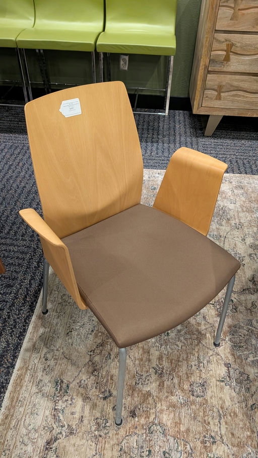 A modern chair features a light wood back, brown seat cushion, and metal legs. It has armrests and is set against a patterned rug and green stools.